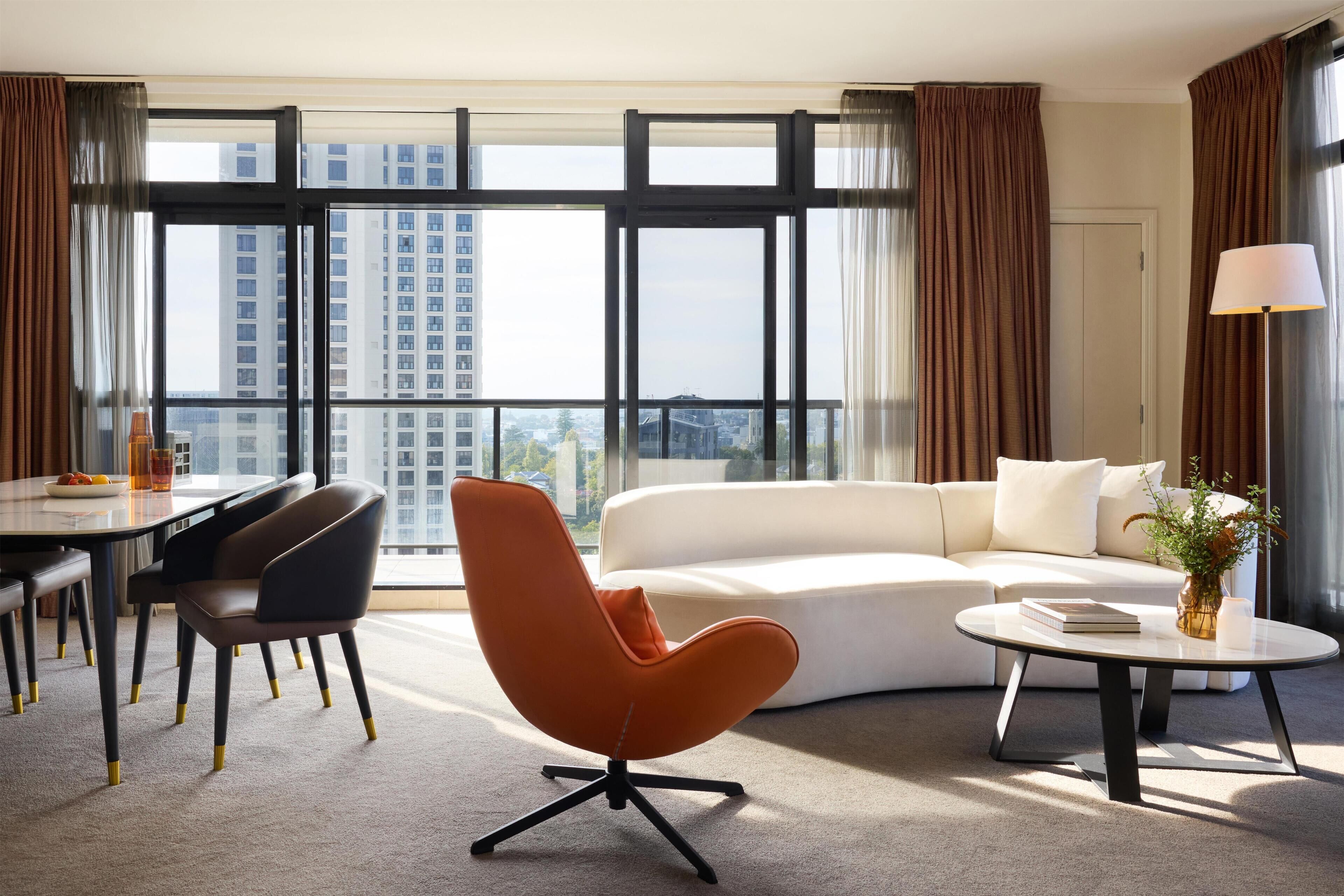 Una habitación de hotel moderna con un ventanal grande, un sofá blanco, un sillón naranja, una mesa de comedor y una lámpara.