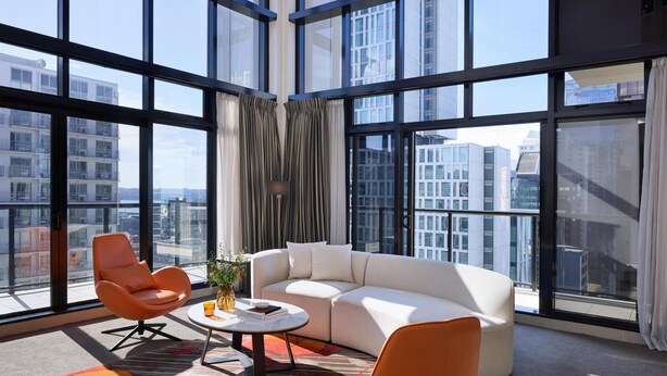 A modern living room with a white sofa, orange armchairs, a coffee table, and large windows offering city views. - Adina Citylife Auckland (Auckland)