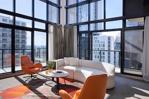 A modern living room with a white sofa, orange armchairs, a coffee table, and large windows offering city views.