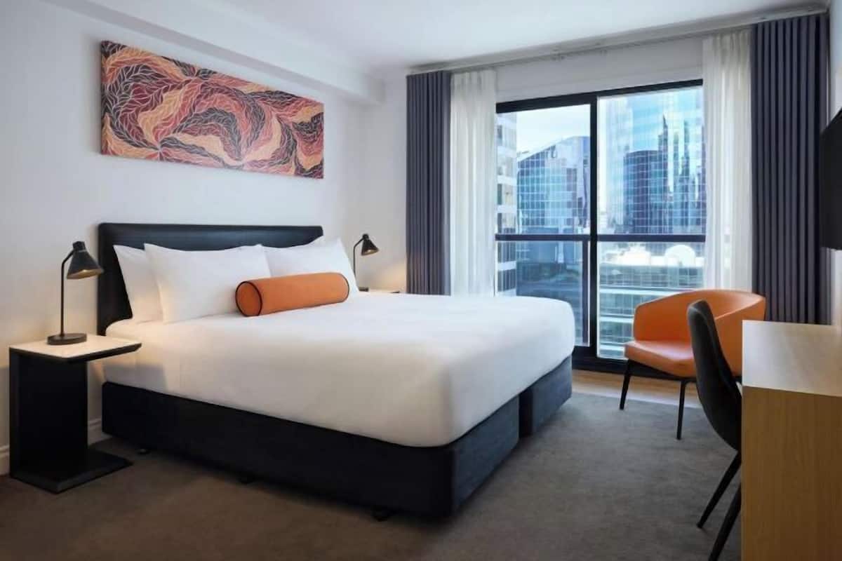 A hotel room with a large bed, a desk, a chair, a window with a city view, and a painting on the wall.