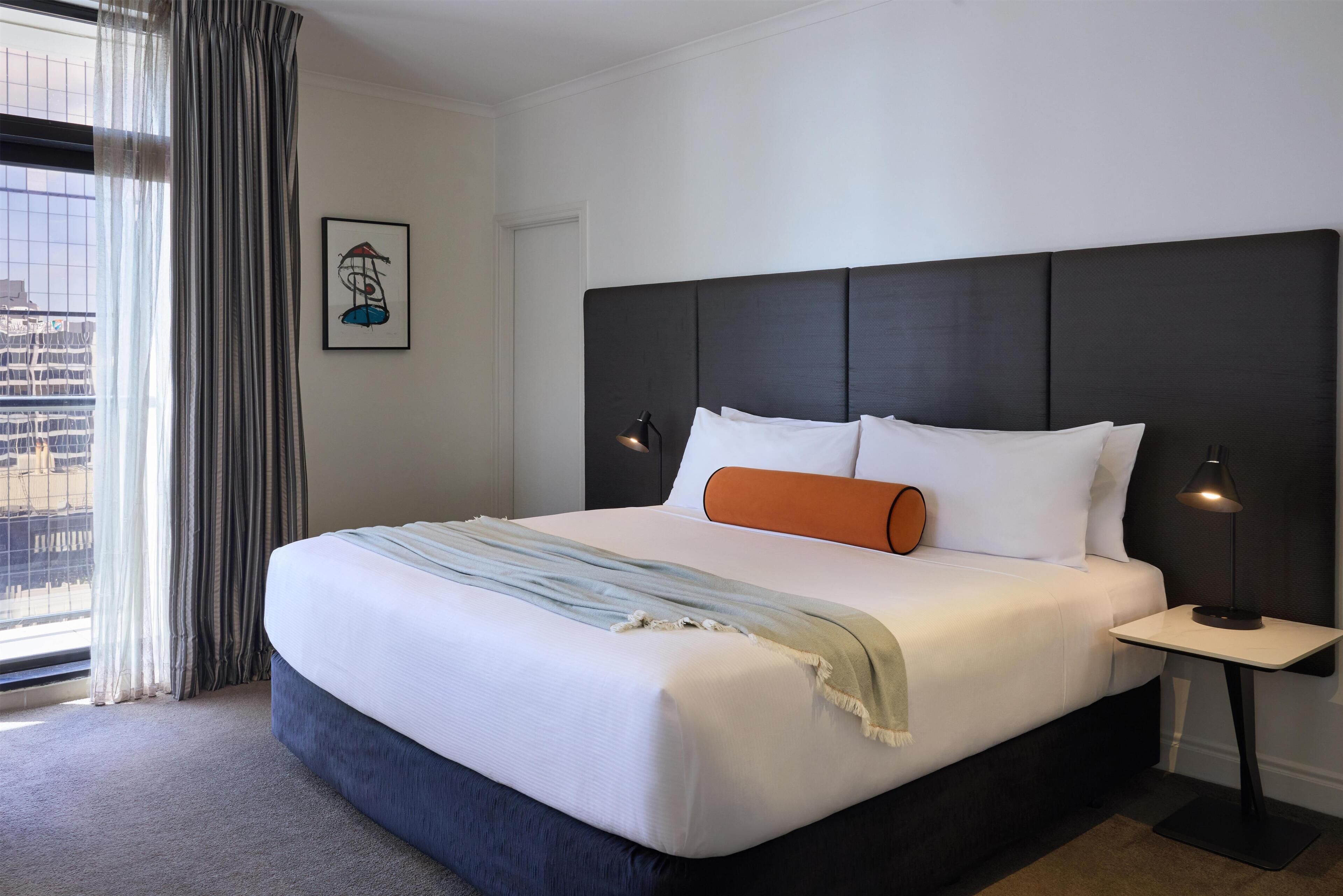 A hotel room with a large bed, a nightstand, a framed picture on the wall, and a view through the window.