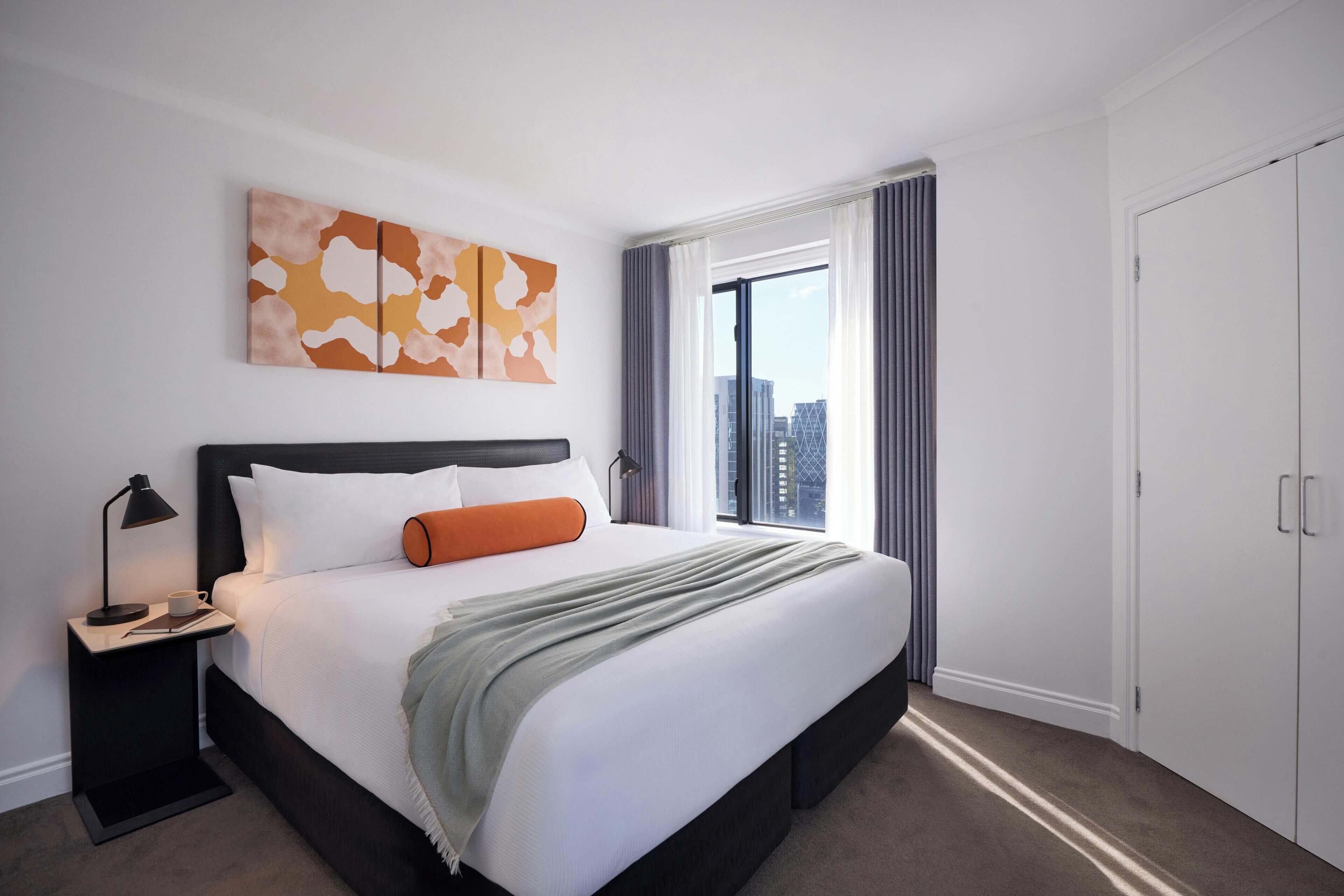 A modern hotel room with a large bed, a nightstand, a window with curtains, and a city view.