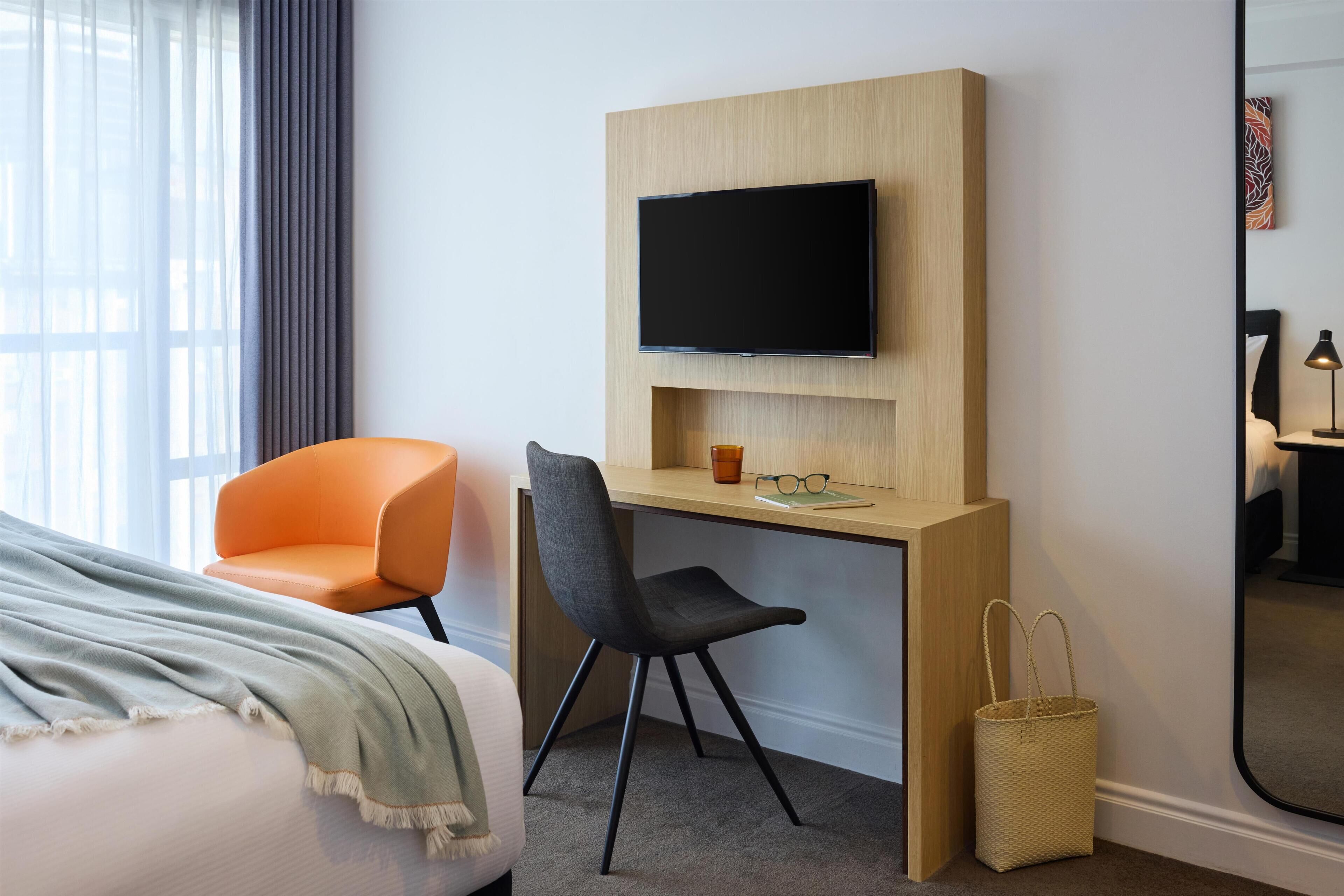 Habitación de hotel con cama, escritorio con televisor, dos sillas, espejo y ventana con cortinas.