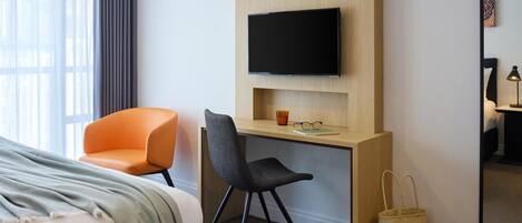A hotel room with a bed, a desk with a TV, two chairs, a mirror, and a window with curtains.