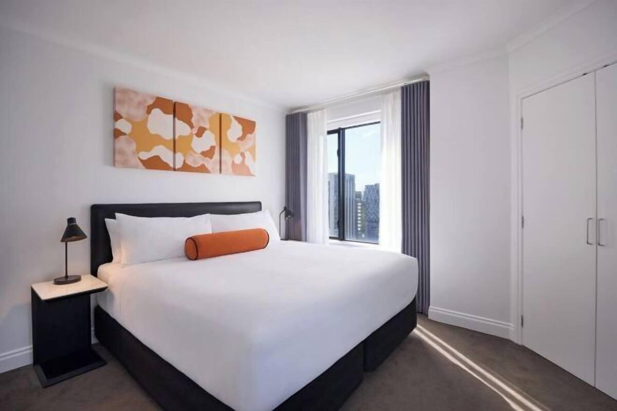 A hotel room with a large bed, a nightstand, a window with curtains, and a view of the city.