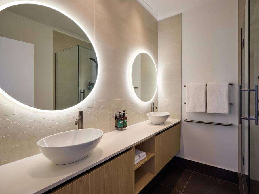Un baño moderno con un espejo ovalado grande, dos lavamanos y una cabina de ducha de vidrio.