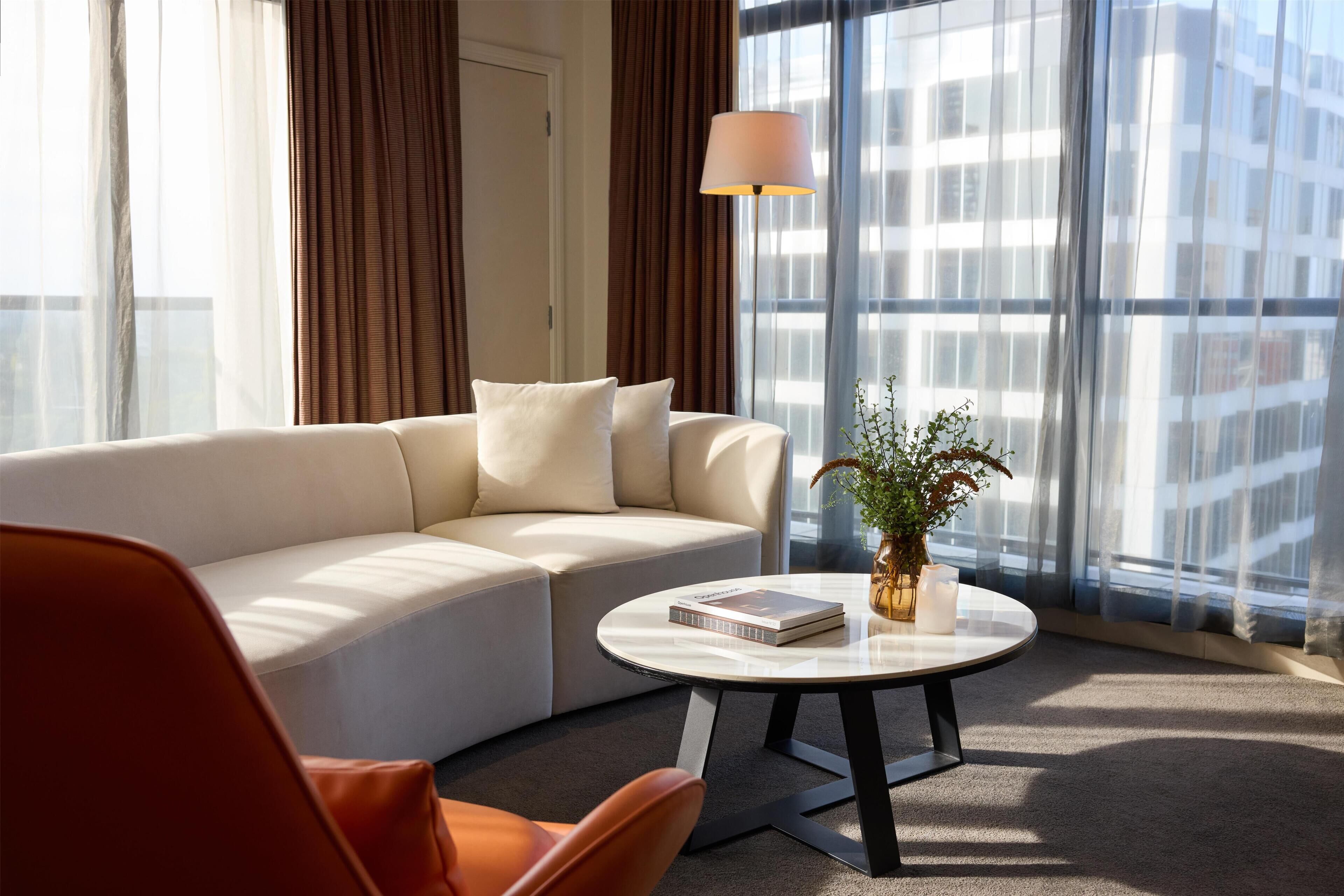 A modern living room with a sofa, a coffee table, a lamp, and a view of a cityscape through large windows.