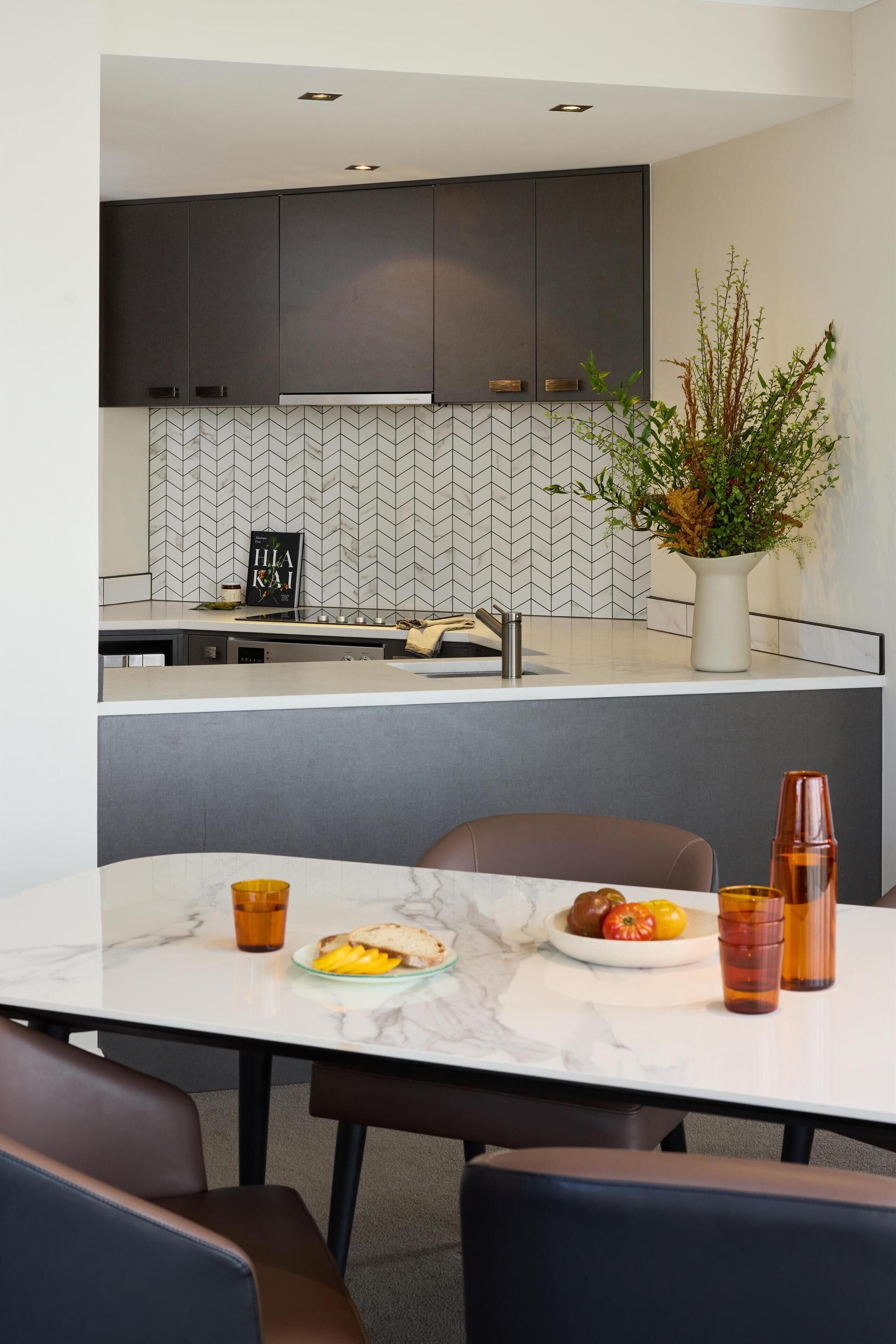Cozinha moderna com armários escuros, backsplash de azulejos em padrão herringbone e área de jantar com mesa e cadeiras de mármore.