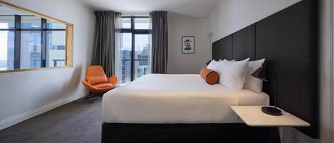 A hotel room with a large bed, a chair, a bedside table, a window with curtains, and a city view.