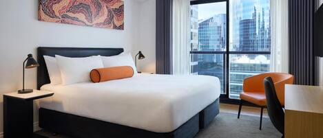 Habitación de hotel con una cama grande, un escritorio, una silla, una mesita de noche, una lámpara, una ventana con vista a la ciudad y una obra de arte abstracta en la pared.