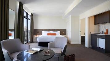 Ein Hotelzimmer mit einem Bett, zwei Sesseln, einem kleinen Tisch, einer Küchenzeile und Blick durch große Fenster.