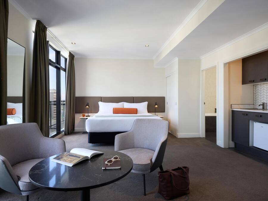 Ein Hotelzimmer mit einem Bett, zwei Sesseln, einem kleinen Tisch, einer Küchenzeile und Blick durch große Fenster.