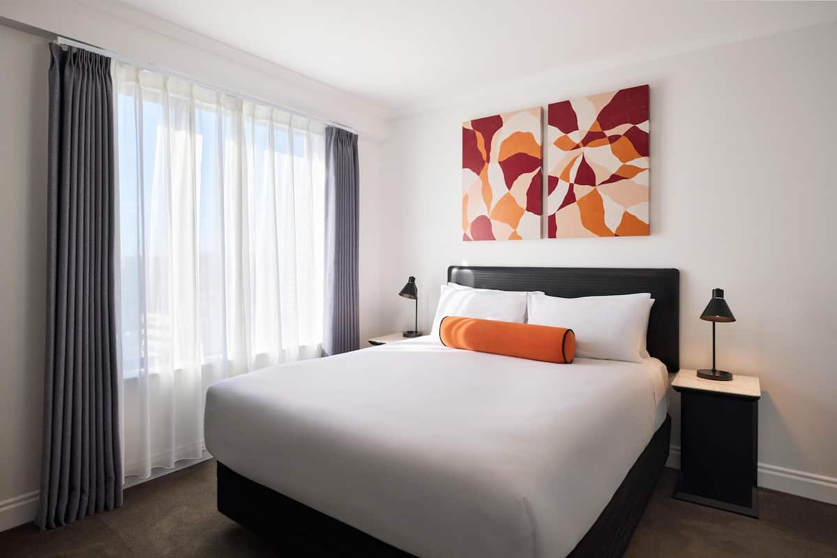 A hotel room with a large bed, two bedside tables with lamps, a window with curtains, and a wall art piece.