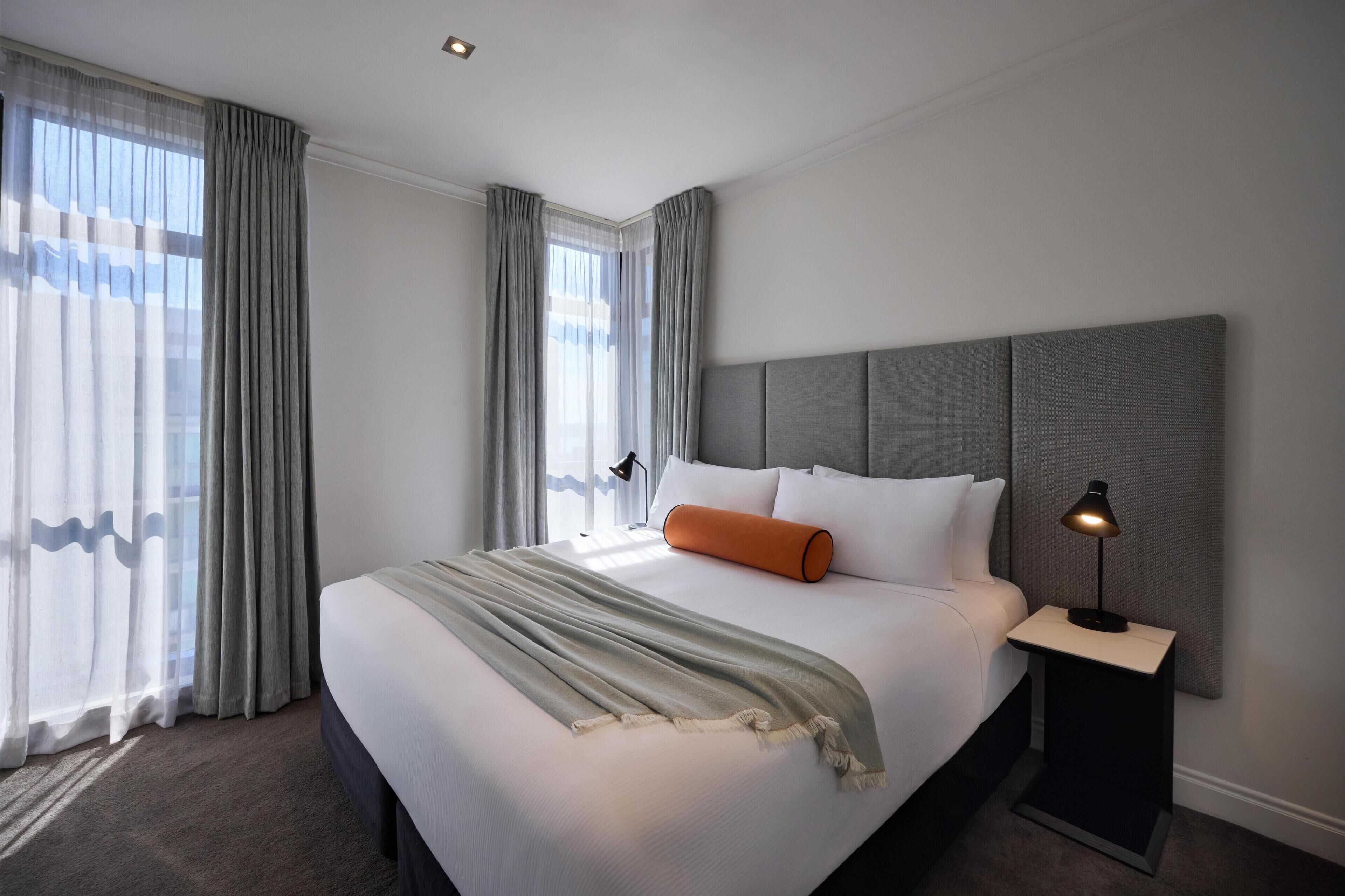 Een netjes opgemaakt bed met een grijze hoofdbord, een bedlamp en een groot raam met gordijnen.