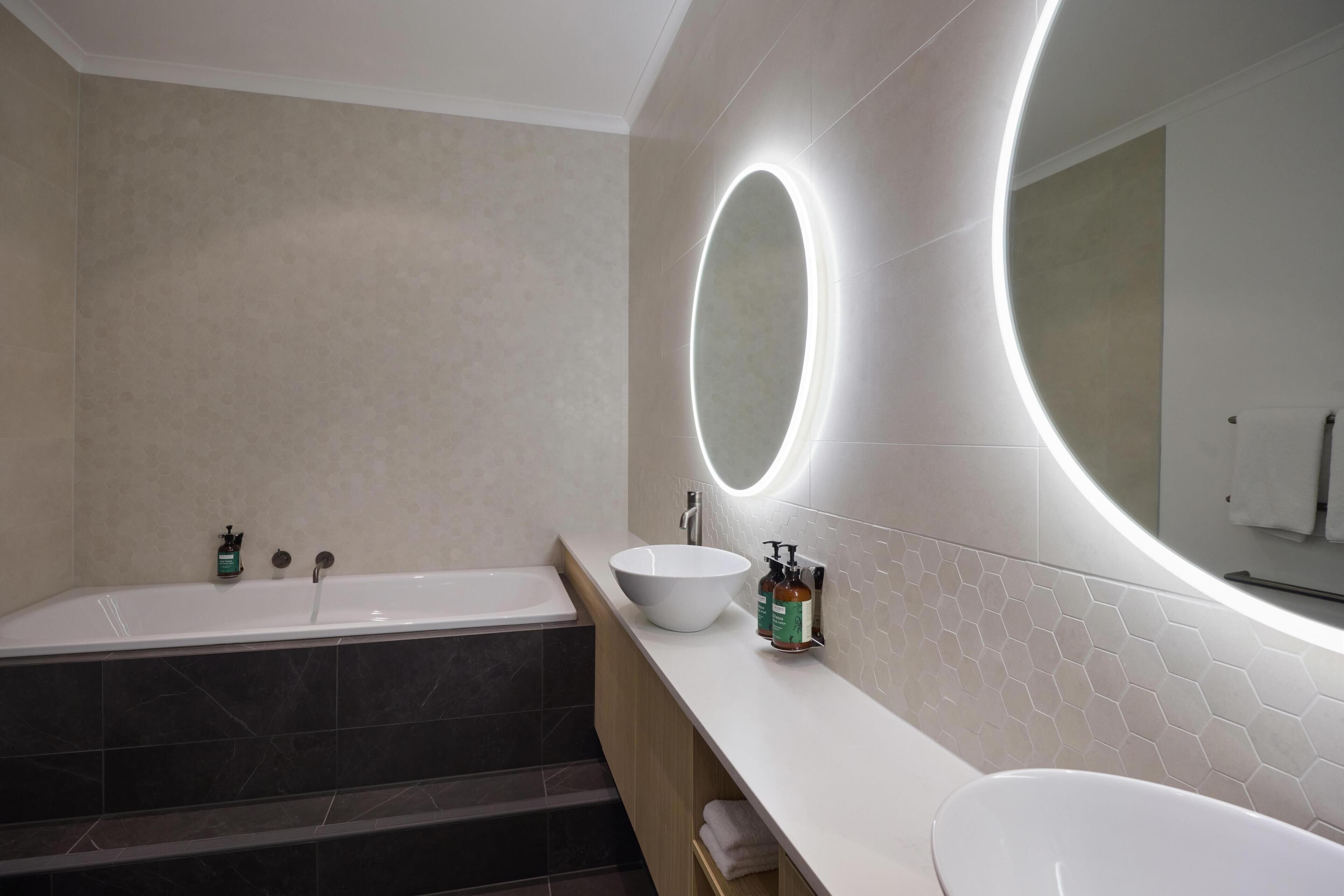 Une salle de bain moderne avec une baignoire sur pieds, un grand miroir rond doté d’un éclairage intégré et un lavabo blanc agrémenté d’un pare-baignoire en carreaux hexagonaux.