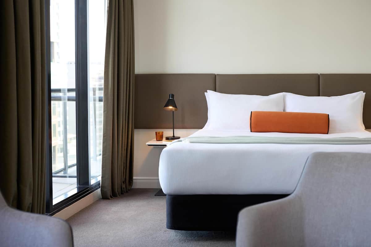 A hotel room with a large bed, a nightstand, a lamp, and a view of the city through the window.