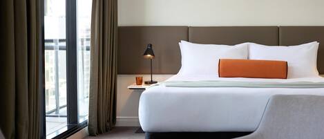 Ein Hotelzimmer mit einem großen Bett, einem Nachttisch, einer Lampe und Blick auf die Stadt durch das Fenster.