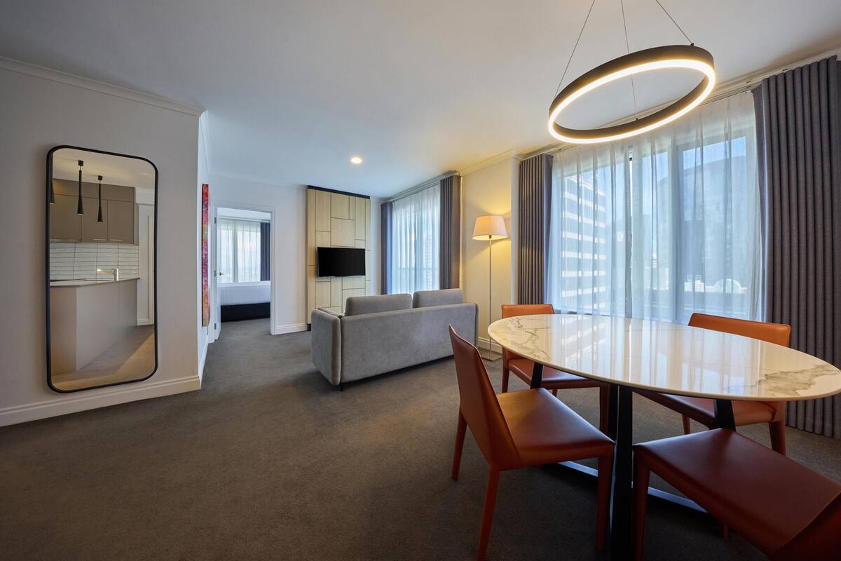 A hotel room with a round marble table, orange chairs, a grey sofa, a flat-screen TV, and a large window with curtains.