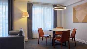 Quarto de hotel com sofá, mesa de jantar com cadeiras, luminária de chão e pintura na parede.