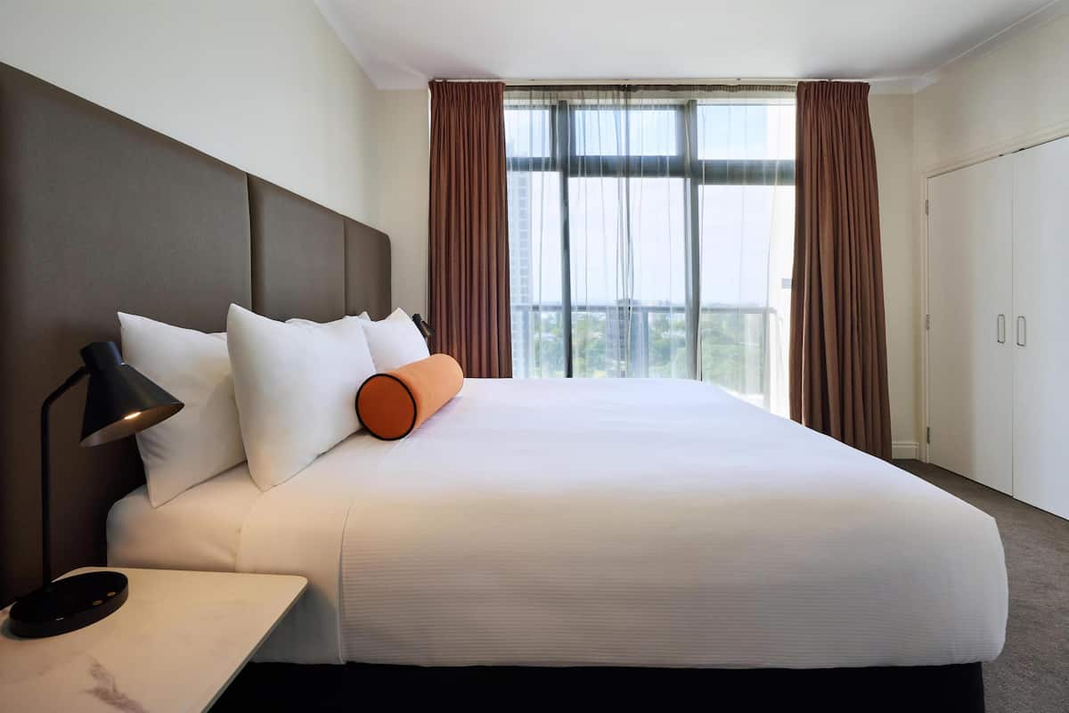 A hotel room with a large bed, a bedside table with a lamp, a window with curtains, and a wardrobe.