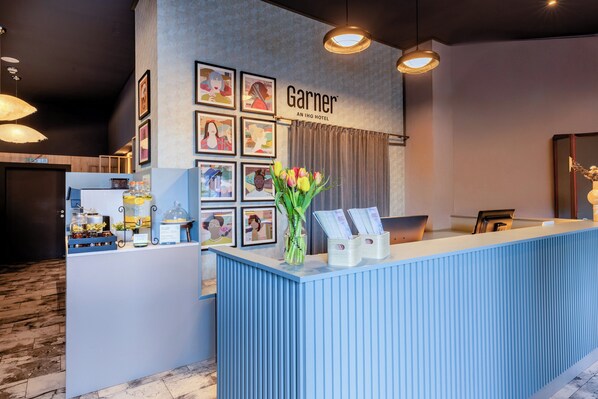 A hotel reception area with a blue counter, framed artwork on the wall, and a vase of flowers. - Garner Hotel Vienna by IHG (Vienna)