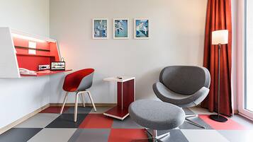Habitación de hotel moderna con una decoración en tonos rojo y gris, que incluye un escritorio, dos sillas, una mesita pequeña y tres cuadros en la pared.