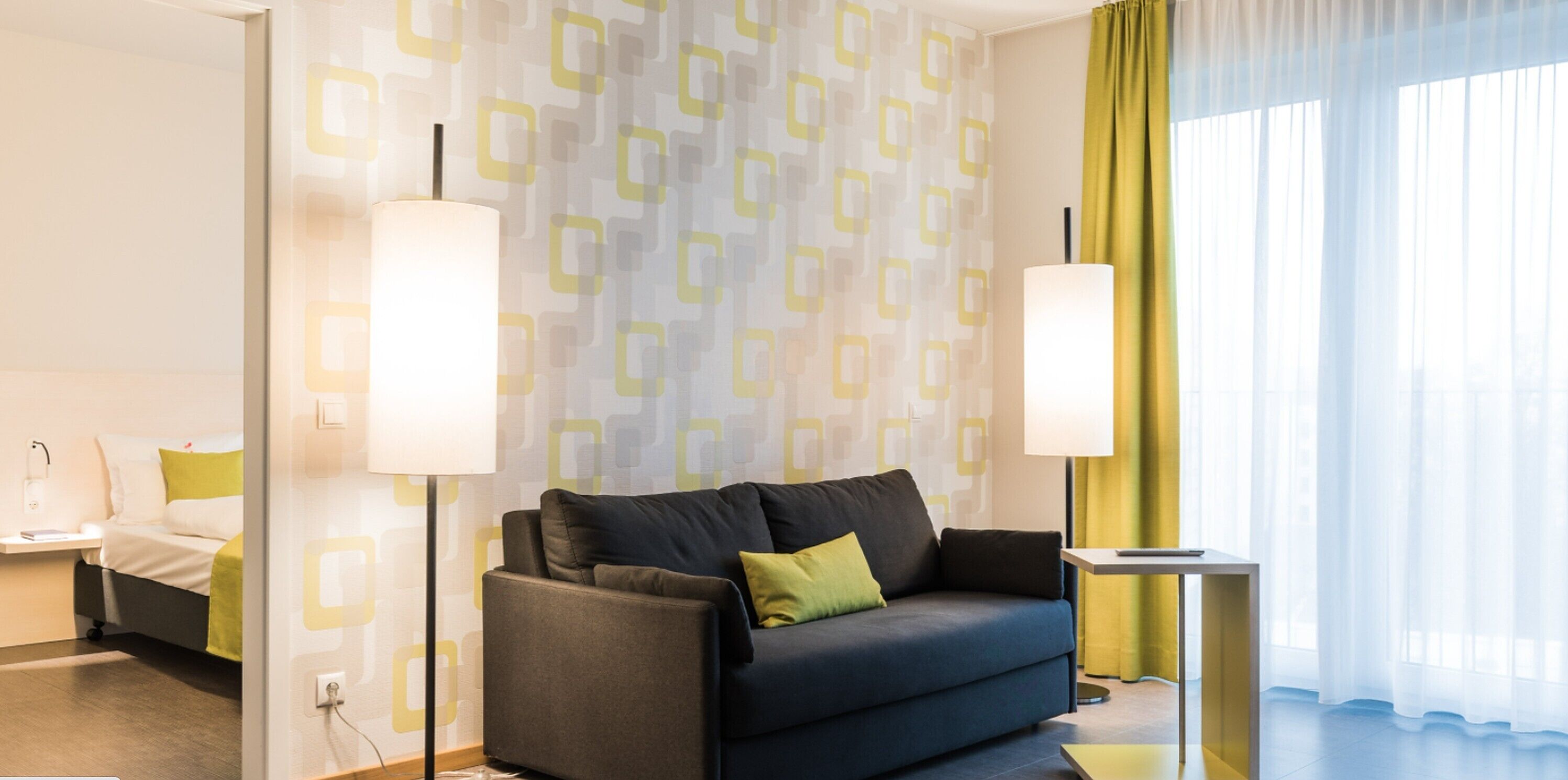 Una habitación de hotel moderna con un sofá azul oscuro, una mesita de noche, una lámpara y una ventana con cortinas translúcidas.