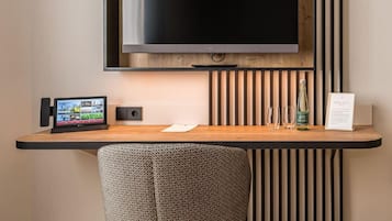 Et moderne værelse med et stort, fladskærms TV monteret på en trævæg, et skrivebord med en bærbar computer, en højttaler og et glas vand.