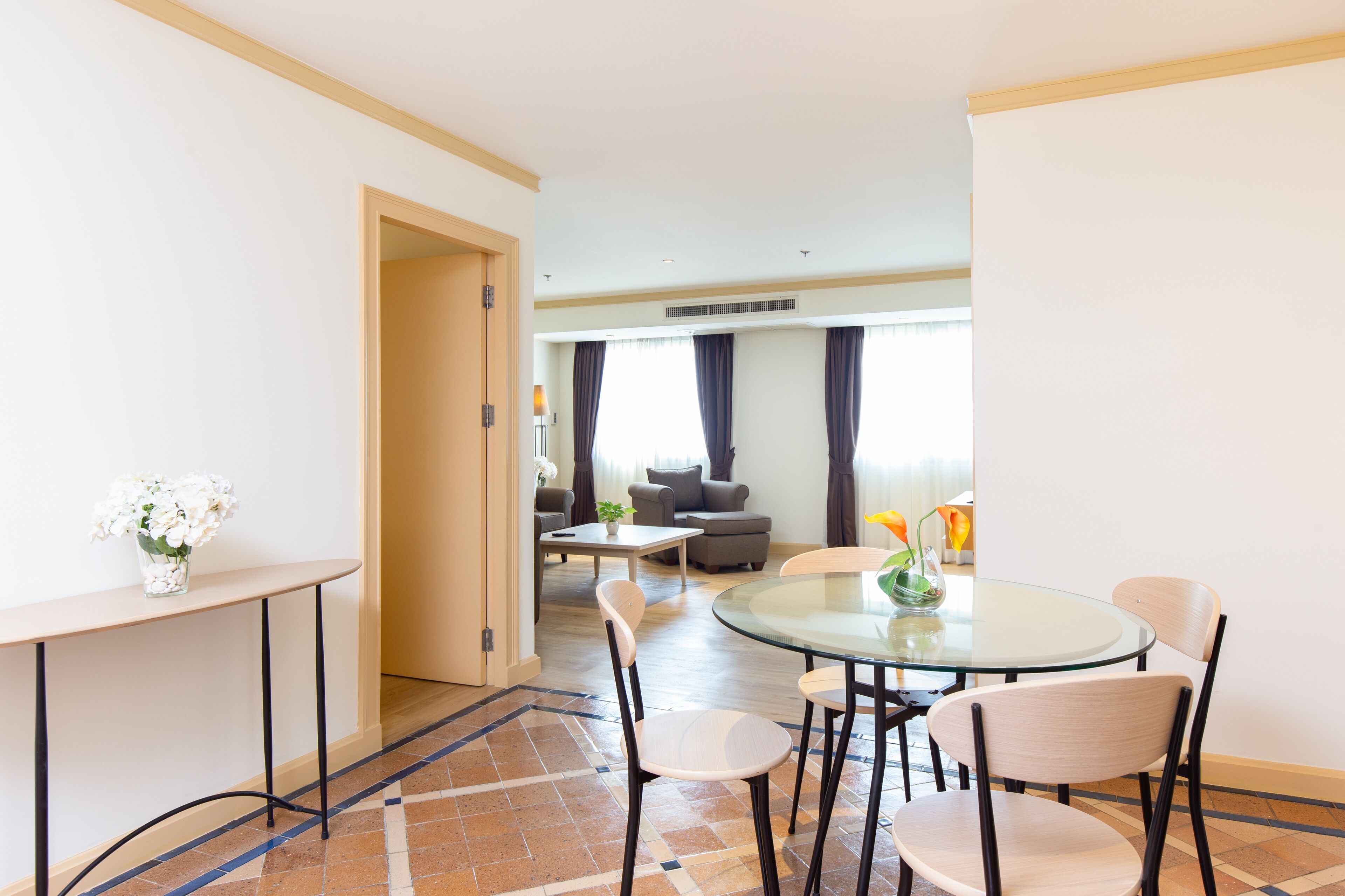 A modern hotel room with a round glass table, four chairs, a small side table with a vase of flowers, and a sofa in the background.