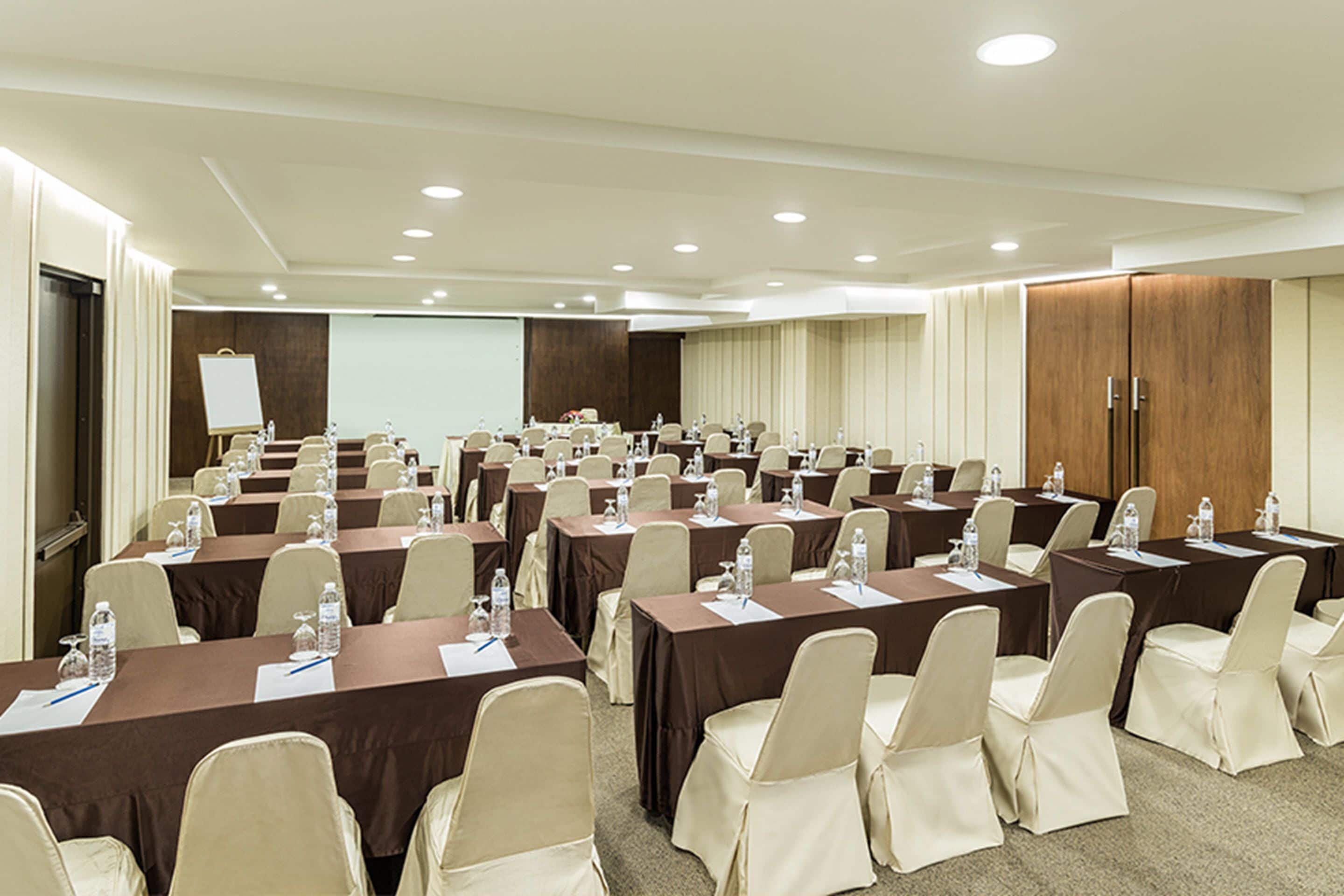 A conference room with multiple tables and chairs, equipped with water bottles and papers, in a well-lit room with a projector screen and wooden cabinets.