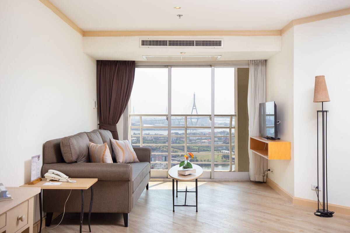 A modern living room with a sofa, a small table, a TV on a shelf, and a large window with a view of a bridge.