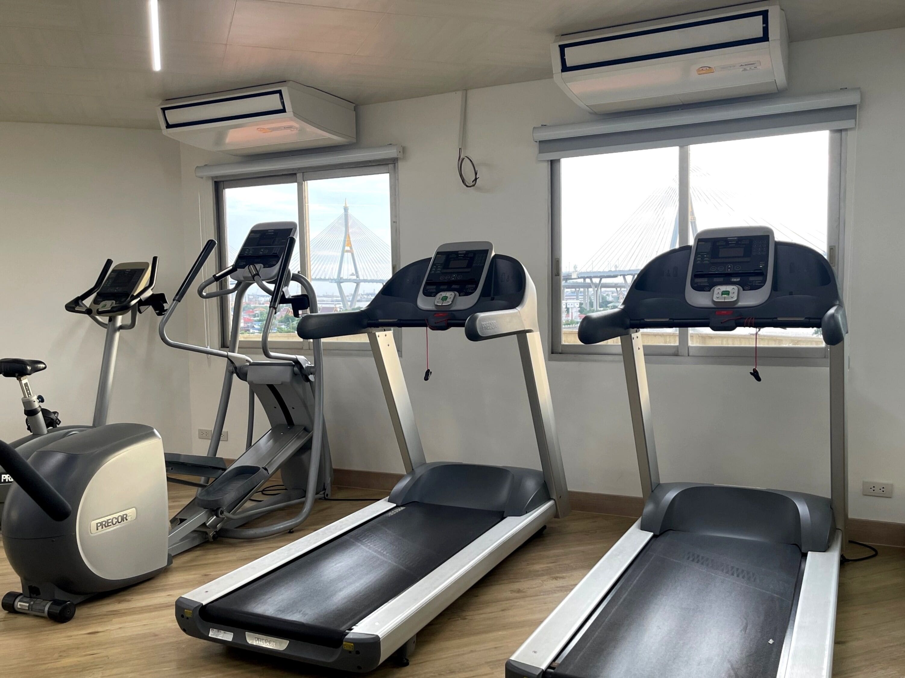 A gym with treadmills, an elliptical, and a stationary bike, large windows with a view of a bridge, and air conditioning units.