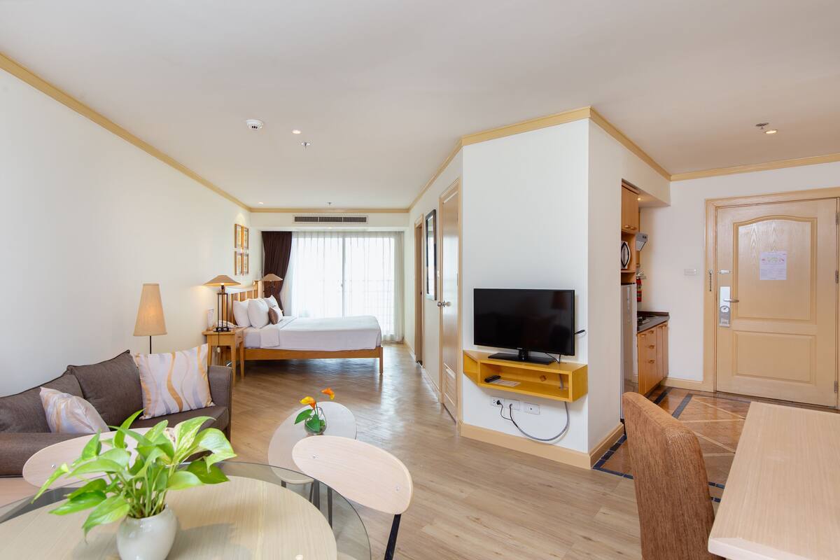 A hotel room with a bed, a TV, a sofa, a dining table, and a small plant.