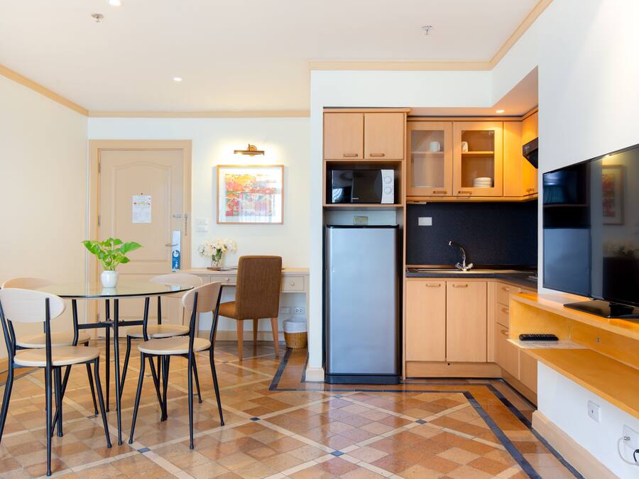A compact kitchen with a refrigerator, microwave, and a flat-screen TV. There is a dining area with a table and chairs, and a small potted plant on the table.