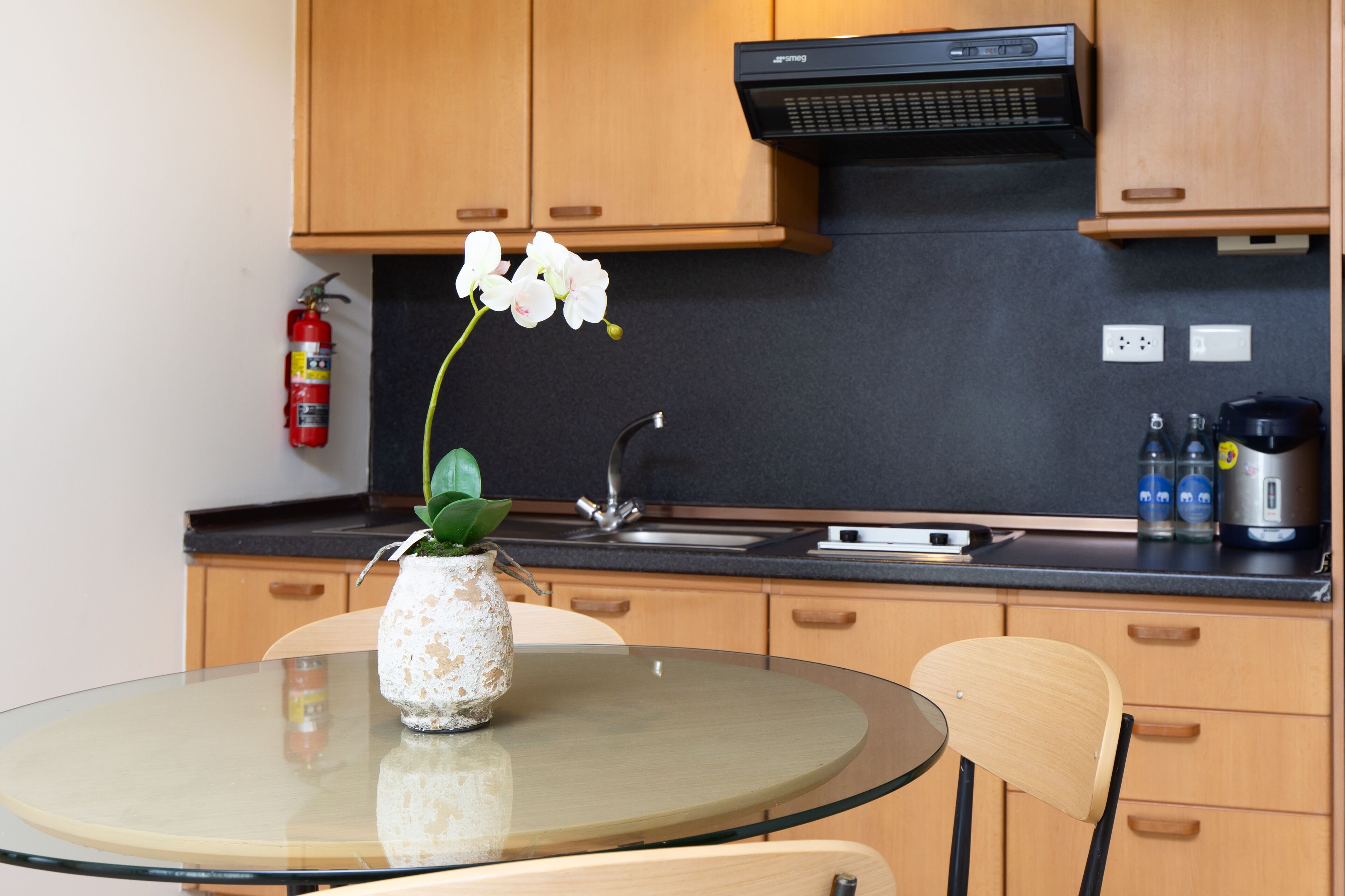 Een moderne keuken met houten kastjes, een zwart aanrecht, een glazen tafel en een vaas met een witte orchidee.