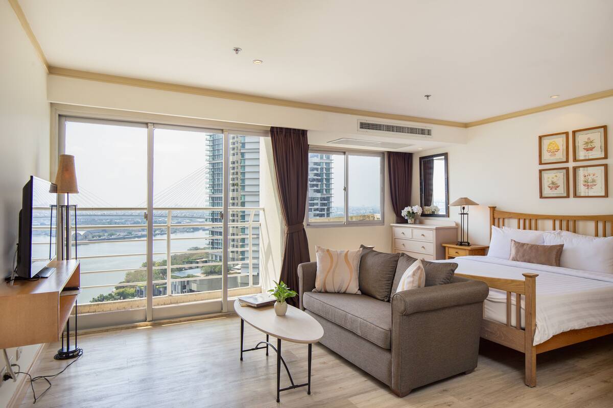 A modern hotel room with a large window, a sofa, a bed, a bedside table, and a TV.