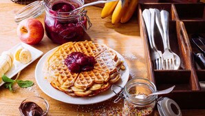 Een bord met wafels, met een kers erop, omringd door appels, een pot jam, een bak chocoladesaus en een potje sprinkles op een houten tafel.