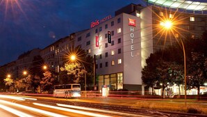 Een modern hotelgebouw met een opvallend 'ibis HOTEL' bord, veel ramen en een bushalte aan de aangrenzende weg.