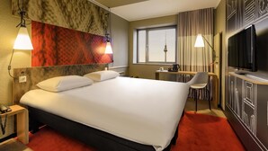 Een hotelkamer met een groot bed, bedlampen, een bureau met stoel, een televisie en uitzicht op een toren door het raam.