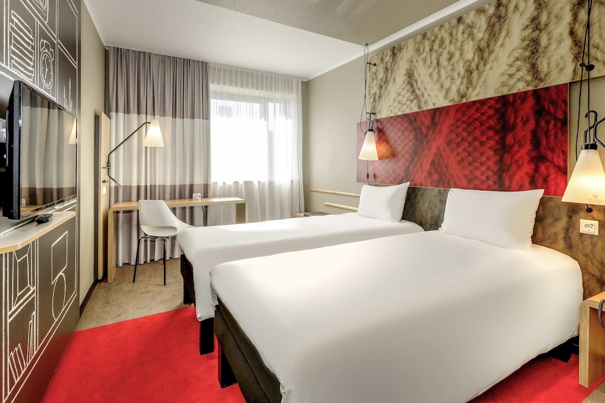 Habitación de hotel con dos camas, un escritorio, una silla, un televisor y una alfombra roja.