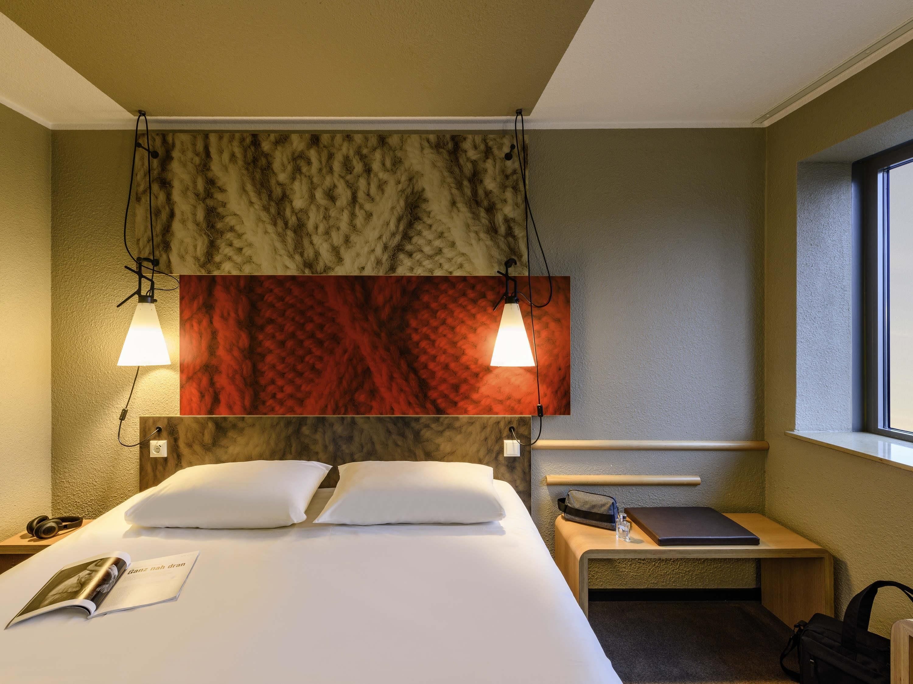 Hotellihuone, jossa on sänky, kaksi yöpöytävalaisinta, yöpöytä, ikkuna ja kuvioitu seinä.