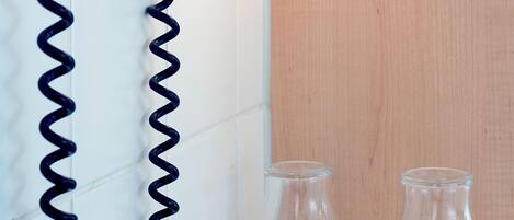 A hair dryer mounted on a wall with a coiled cord, a small control panel, and two empty glasses on a counter.