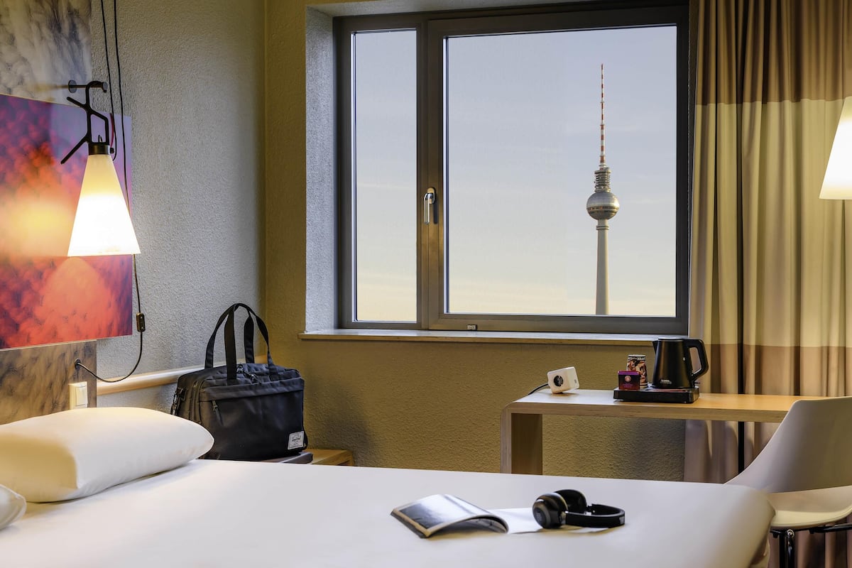 Habitación de hotel con una cama, un escritorio con lámpara, una silla, una ventana con vistas a una torre y una bolsa sobre la cama.