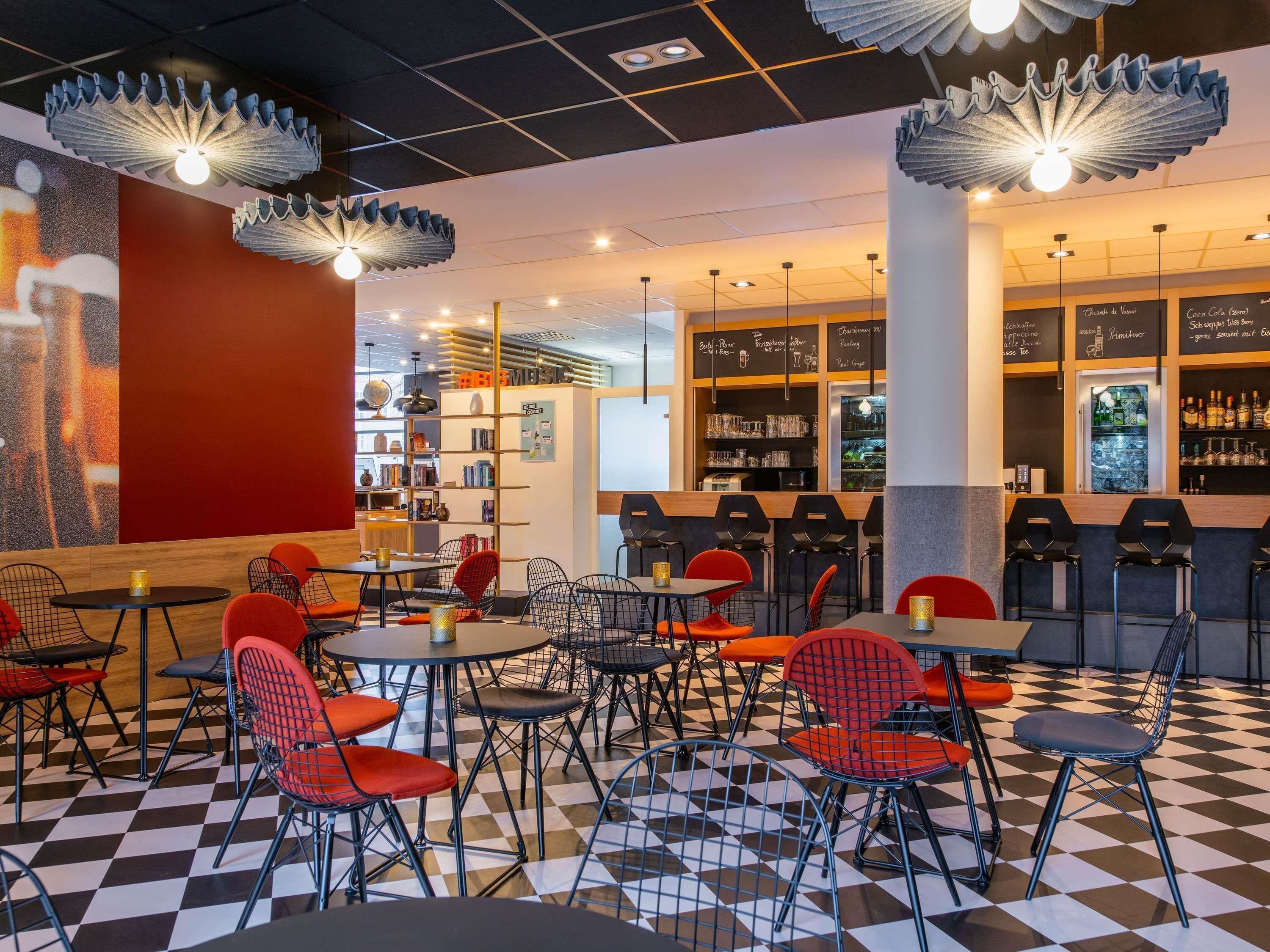 Ett kafé med rutigt golv, röda och svarta stolar, en bardisk med hyllor fulla av drycker och en vägg med en stor, röd sektion.