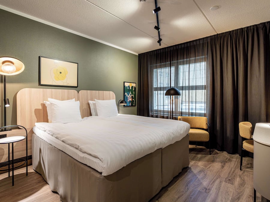 Une chambre d’hôtel avec un grand lit, un bureau, une chaise, une lampe et une fenêtre avec des rideaux.