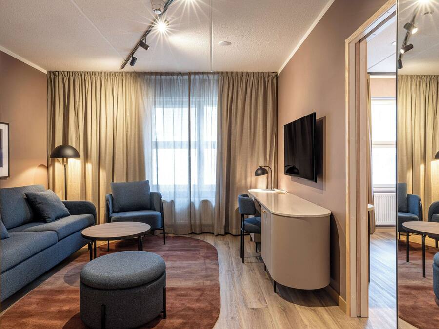 Una habitación de hotel moderna con sofá, sillón, reposapiés y un pequeño escritorio con silla. Hay un televisor de pantalla plana montado en la pared y un espejo grande que refleja la habitación.