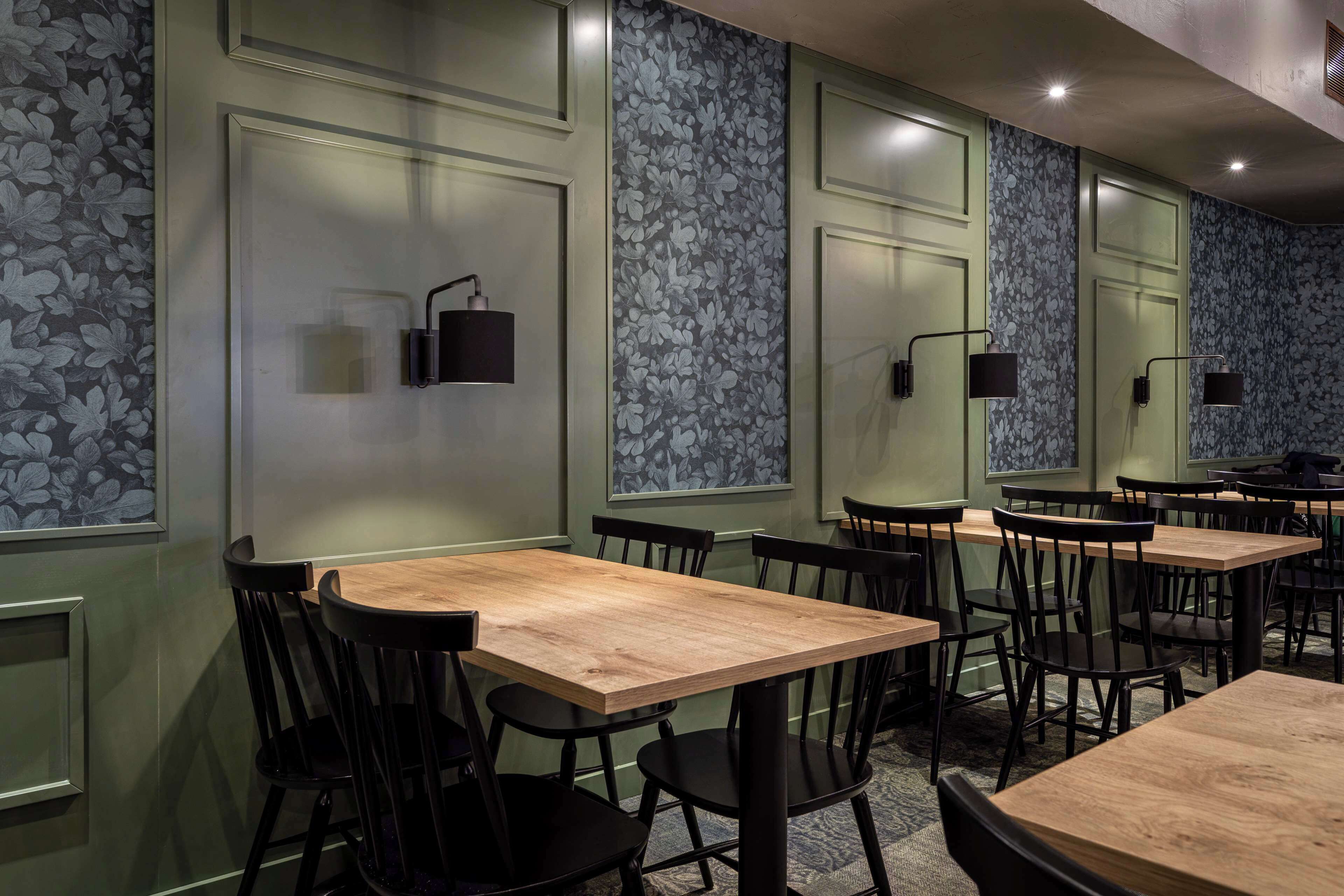 A restaurant with wooden tables and black chairs, green walls with floral wallpaper, and black pendant lights.