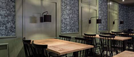 Um restaurante com mesas de madeira e cadeiras pretas, paredes verdes com papel de parede floral e luminárias pendentes pretas.