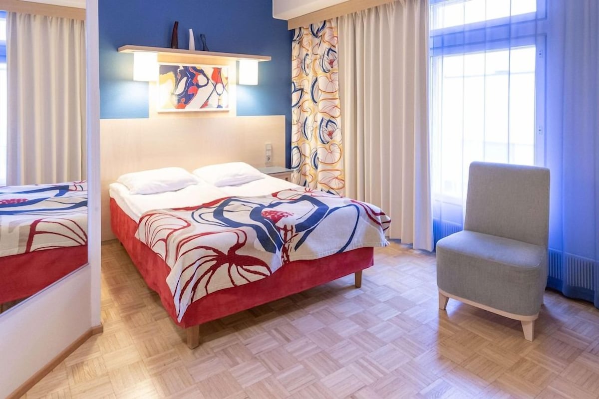 Habitación de hotel con cama, una silla, estante en la pared y ventana con cortinas.