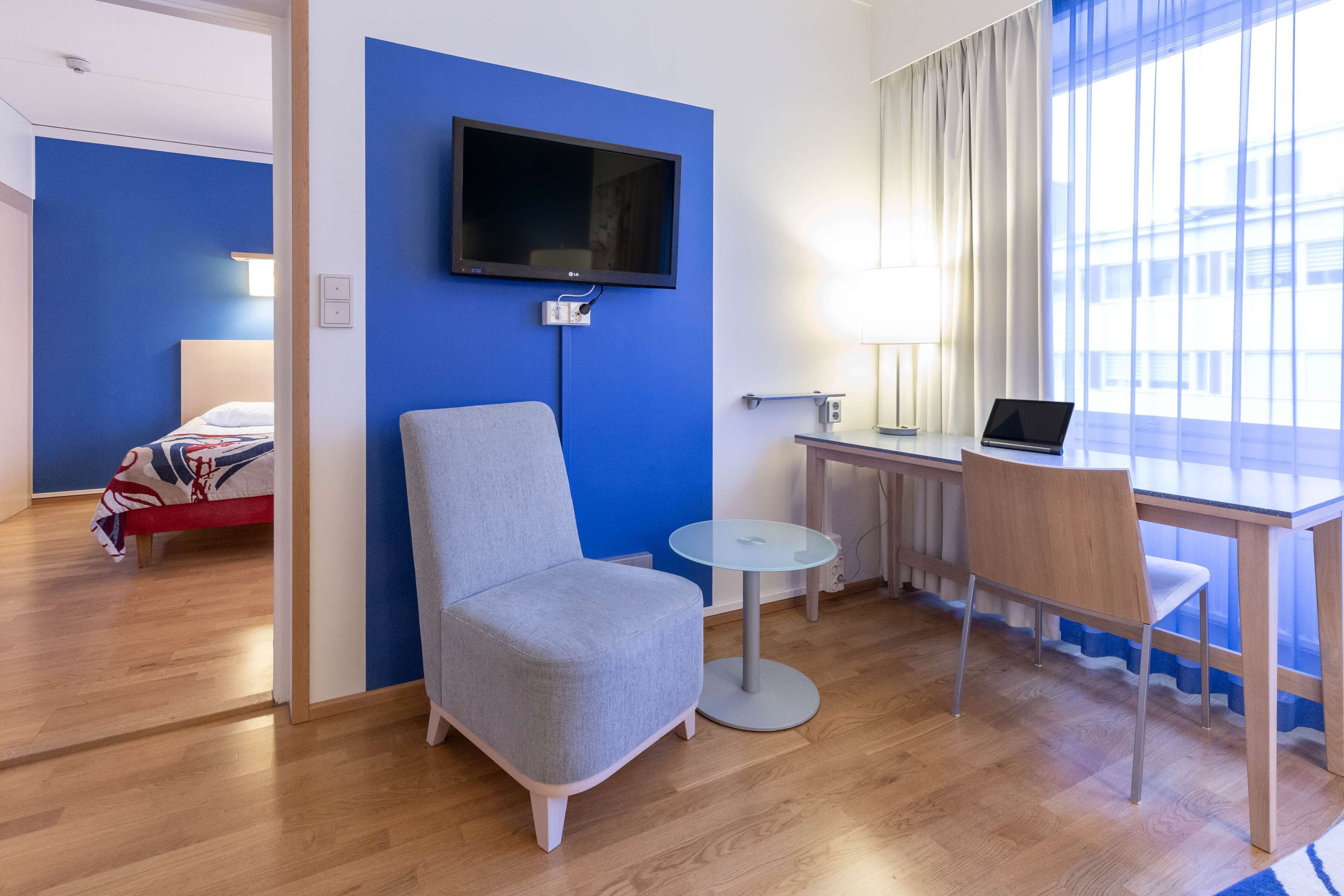 Ein Hotelzimmer mit blauer Akzentwand, einem Flachbildfernseher, einem Schreibtisch mit einem Laptop, einem grauen Sessel und einem Bett mit einer rot-weißen Decke.
